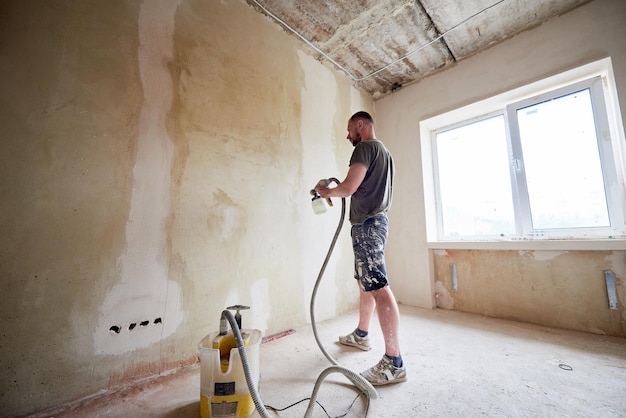 Homme peignant un mur gris avec un pistolet pulvérisateur rénovant une nouvelle maison Récipient ouvert avec une peinture