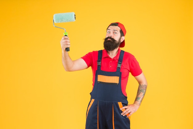 Homme peindre le constructeur de hipster mur en casquette fond jaune assistant monteur en uniforme de travail ingénieur concepteur prêt à peindre le concept de réparation de mur ouvrier barbu avec outil à rouleaux