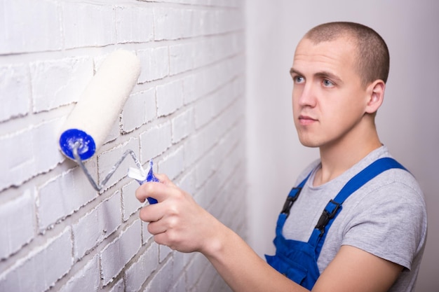 Homme peintre en vêtements de travail peignant un mur de briques avec un rouleau à peinture