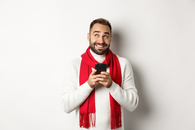 Homme pensant tout en envoyant un message texte sur téléphone mobile, souriant et debout sur fond blanc