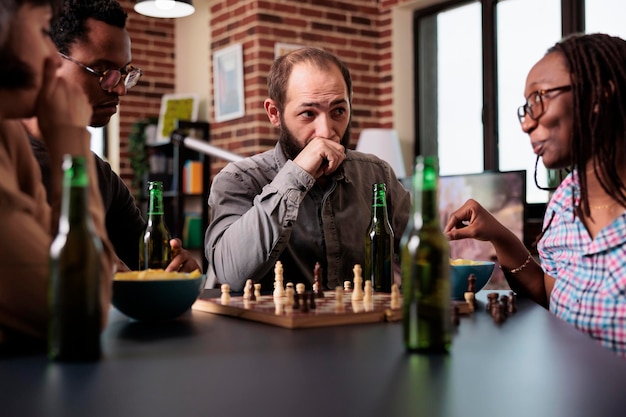 Homme pensif assis à table dans le salon avec des amis multiethniques tout en jouant aux échecs. Personne tendue appréciant les jeux de société de stratégie tout en pensant au prochain mouvement de l'échiquier.