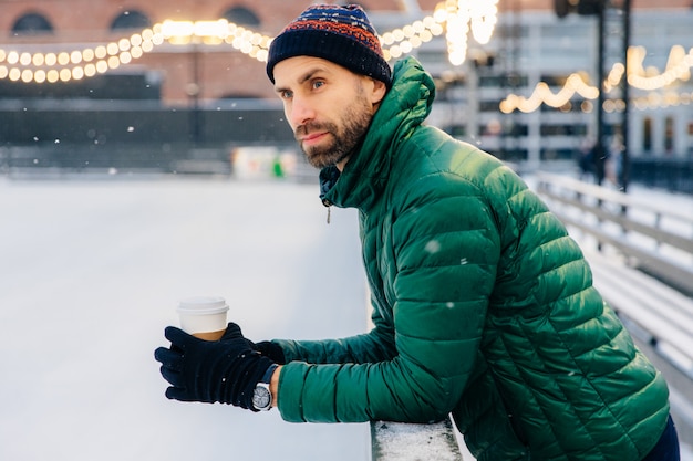 Un homme pensif, à la barbe épaisse, se penche, tient du café à emporter, regarde un match de hockey en hiver
