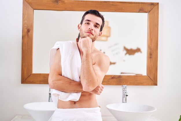 L'homme pensif avec une serviette sur l'épaule se tient près du miroir dans le bain le matin