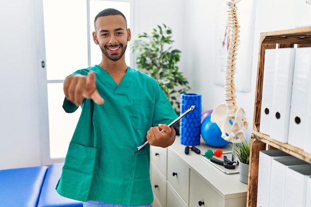 Homme physiothérapeute afro-américain travaillant à la clinique de récupération de la douleur pointant vers vous et la caméra avec les doigts, souriant positif et joyeux