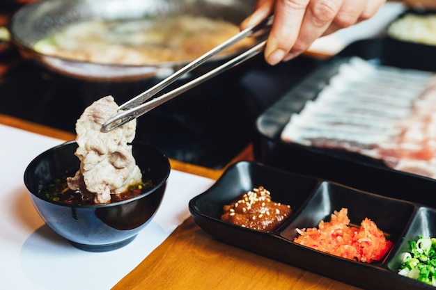 Un homme pince le porc Kurobuta bien cuit et le trempe dans une sauce ponzu avec une pince.