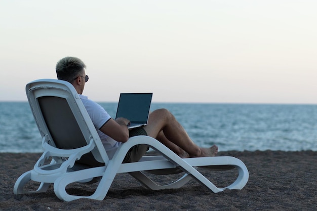 Un homme sur la plage avec un ordinateur portable sur un fauteuil de salon travaillant le concept de travailler en ligne n'importe où dans le monde