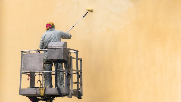 Homme Sur Une Plate-forme élévatrice Peignant Le Mur Du Bâtiment Avec Un Extérieur Extérieur De Rouleau.