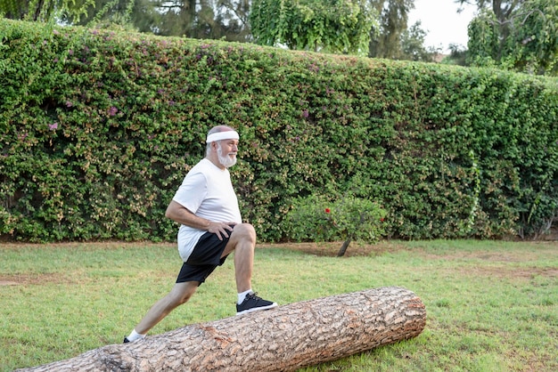Homme plus âgé faisant un squat avant sur une jambe vers l'avant dans le parc de la ville pendant l'entraînement de routine du matin s'appuyant sur le tronc de l'arbre