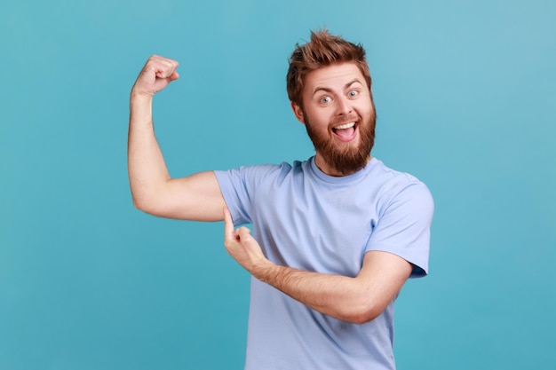 Homme pointant le doigt sur son muscle du bras pompant dans la remise de gym regardant la caméra avec un visage satisfait