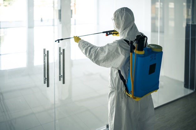 Un Homme Portant Une Combinaison De Désinfection Pulvérisant De Désinfectant Sur Les Poignées Des Portes En Verre Dans Un Centre Commercial Vide Pour Empêcher La Propagation Du Covid-19. Sensibilisation à La Santé, Concept Propre, Défense.