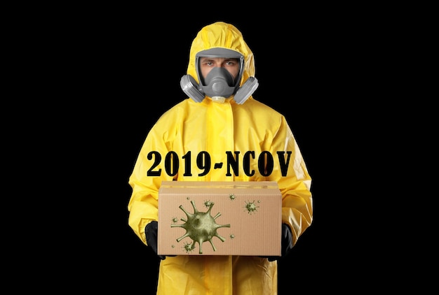 Homme portant une combinaison de protection chimique avec boîte en carton sur fond noir Éclosion de coronavirus