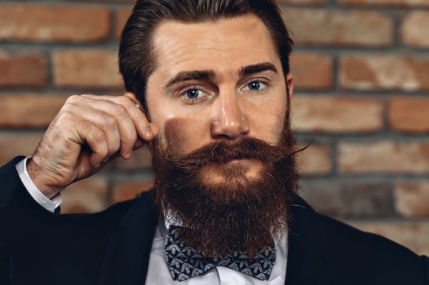 Photo homme portant un costume, une moustache et une barbe de hussard et posant contre un mur de briques et touchant sa moustache.beauté