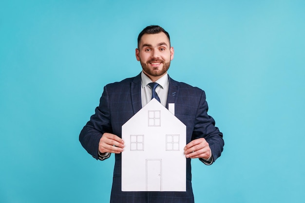 Photo homme portant un costume de style officiel tenant une maison en papier avec des fenêtres et des portes dessinées faisant le projet de la maison de ses rêves en regardant la caméra avec le sourire studio intérieur tourné isolé sur fond bleu