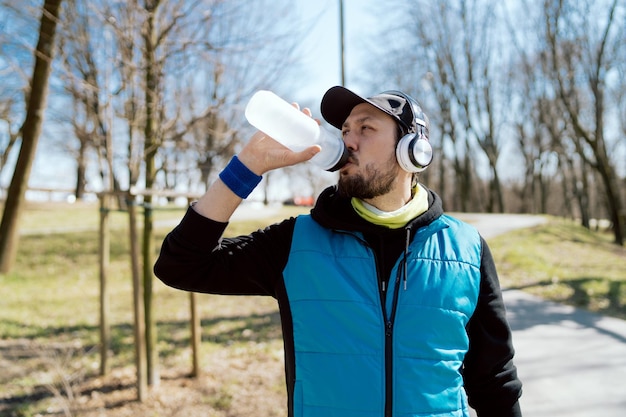 Un homme portant des écouteurs sans fil boit de l'eau d'une bouteille réutilisable un athlète fait du jogging dans le parc