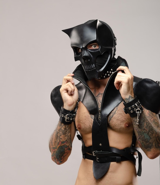 Un homme portant un masque de crâne de démon bdsm vêtu d'une cape en cuir avec des bracelets et des lanières en cuir sur son corps