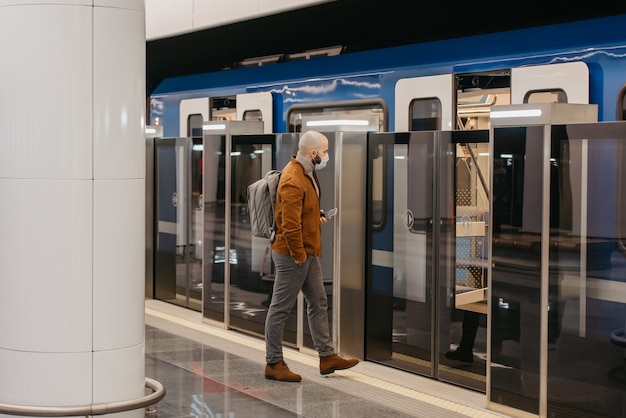 Un homme portant un masque médical pour éviter la propagation du coronavirus tient un téléphone portable en entrant dans la voiture de métro moderne. Un homme chauve portant un masque chirurgical garde une distance sociale.
