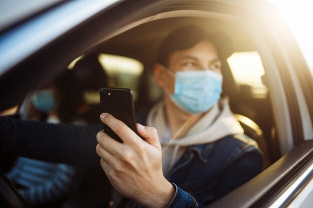 Un homme portant un masque médical tient un téléphone portable dans sa main tout en conduisant une voiture