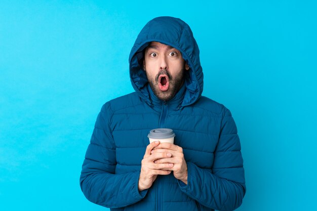 Homme portant une veste d'hiver et tenant un café à emporter sur un mur bleu isolé avec une expression faciale surprise