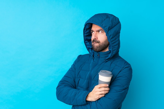 Homme portant une veste d'hiver et tenant un café à emporter sur un mur bleu isolé avec une expression de visage confuse