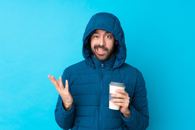 Homme portant une veste d'hiver et tenant un café à emporter sur un mur bleu isolé faisant un geste de doute