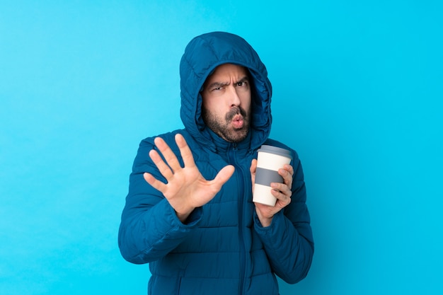 Homme portant une veste d'hiver et tenant un café à emporter sur le mur bleu isolé nerveux qui s'étend les mains vers l'avant