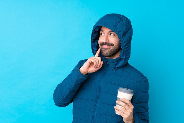 Homme portant une veste d'hiver et tenant un café à emporter sur le mur bleu isolé en pensant à une idée tout en levant