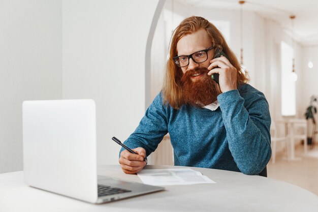 Homme positif aux longs cheveux roux travaillant et parlant au téléphone