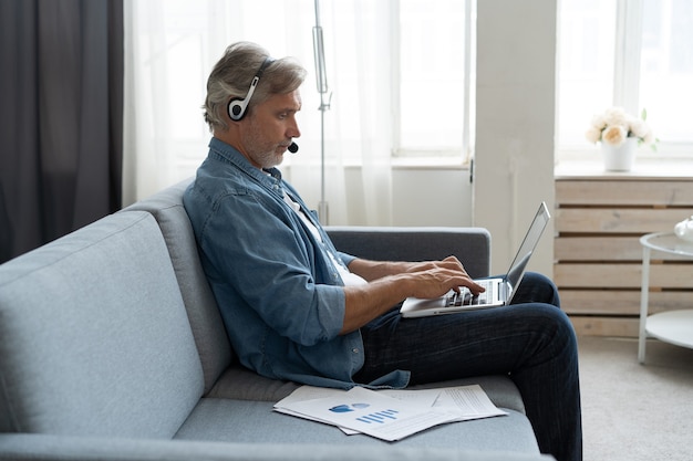 Homme positif dans les écouteurs à l'aide d'un ordinateur portable écoutant un podcast ou prenant part à un webinaire assis sur un canapé à la maison.