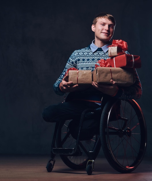 Homme positif en fauteuil roulant avec des cadeaux de Noël sur fond gris foncé.