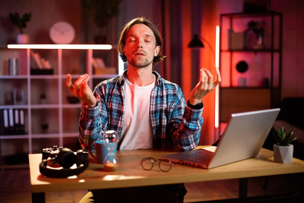 Homme positif méditant les yeux fermés assis au bureau avec un ordinateur portable moderne