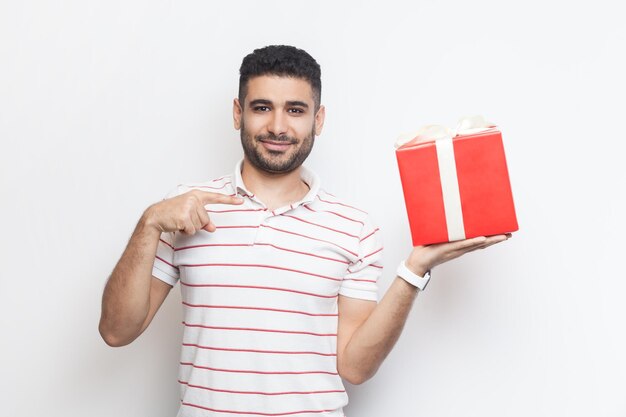 Homme positif souriant portant un t-shirt debout tenant une boîte cadeau enveloppée de rouge pointant vers un cadeau