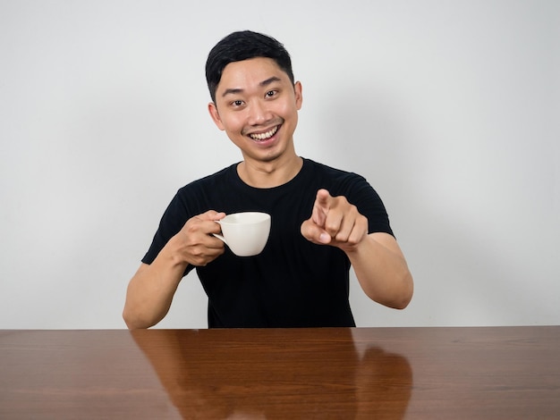 Un homme positif sourit et tient une tasse de café assis à la table pointant du doigt vers vous