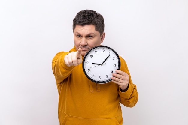 Homme positif tenant une grande horloge murale et pointant vers la caméra avec un sourire agréable, il est temps d'y aller