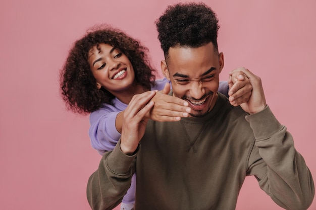 Homme positif en tenue marron riant et tenant les mains de sa copine Mec cool et femme brune frisée vêtue d'une chemise violette souriant sur fond rose