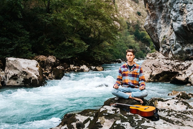 Homme en position méditative avec guitare assis sur la rive d'une rivière de montagne