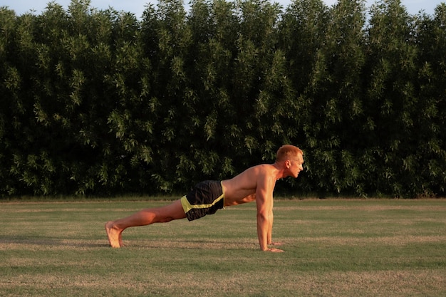 L'homme pratique l'exercice de planche sur les bras droits pour renforcer le noyau sur l'herbe dans le jardin éclairé par la lumière du soleil