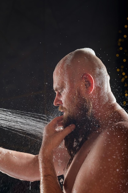 Un homme prend une douche Un homme brutal avec une barbe rousse se tient dans la salle de bain sous l'eau courante et se lave Pulvérisation sur fond noir