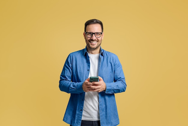 Un homme professionnel souriant envoie des textos sur son téléphone portable tout en se tenant debout avec confiance sur un fond jaune