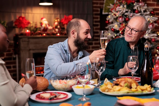 Homme Proposant Des Toasts De Noël, Disant Ses Souhaits Lors D'un Dîner De Fête, Tenant Un Verre De Vin Mousseux Lors D'un Festin à La Maison De Noël. Famille Célébrant Les Vacances D'hiver Ensemble, Buvant à La Fête Du Nouvel An