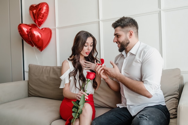 L'homme propose à la femme avec anneau et rose rouge à la maison sur un canapé avec des ballons en forme de coeur