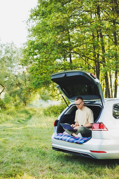 Homme prospère travaillant à l'extérieur de sa voiture assis dans le coffre et regardant l'ordinateur portable Concept de mode de vie de travailleur à distance en ligne Espace de copie