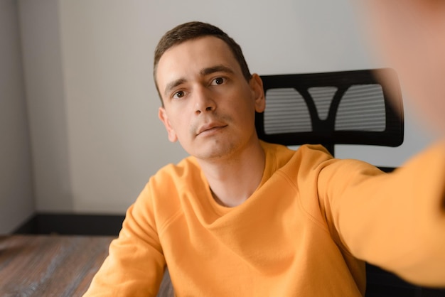 Un homme en pull jaune prend un selfie assis à un bureau à la maison
