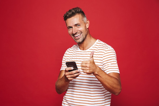 Homme de race blanche 30s en t-shirt rayé souriant et tenant un téléphone portable isolé sur rouge