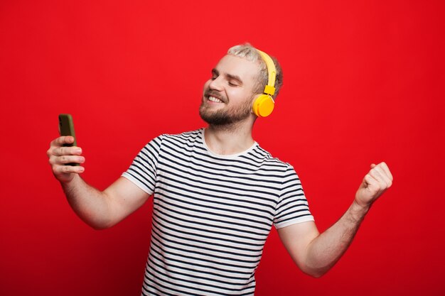 Homme de race blanche aux cheveux blonds dansant sur un mur rouge tout en écoutant de la musique avec des écouteurs