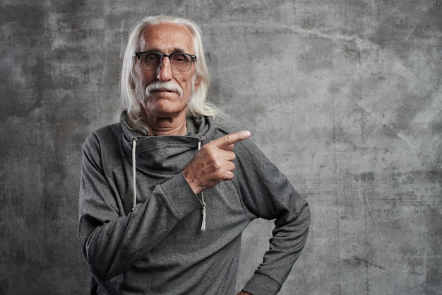 Un homme de race blanche aux cheveux gris âgés pointe sur le côté avec l'index, annonce quelque chose. Grand-père à lunettes avec moustache recommande d'acheter votre produit