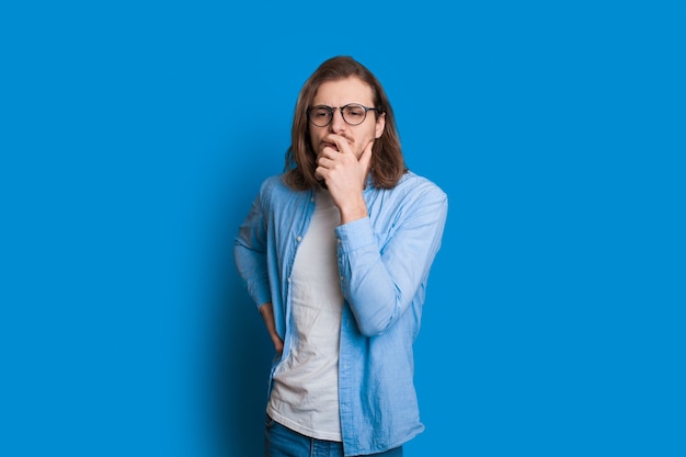 Homme de race blanche aux cheveux longs et belle barbe touche son menton tout en regardant de côté à travers ses lunettes et posant sur fond bleu