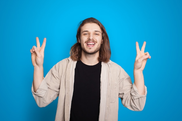 Homme de race blanche aux cheveux longs gesticulant le signe bonjour et pièce sourit sur fond bleu