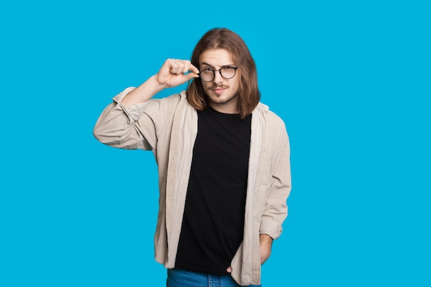 Homme de race blanche aux cheveux longs touchant ses lunettes et regardant la caméra sur un mur bleu