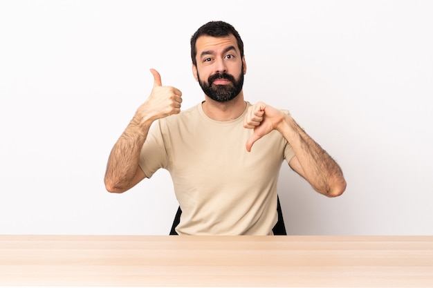 Homme de race blanche avec barbe dans une table faisant bon-mauvais signe. Indécis entre oui ou non.