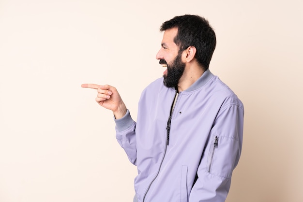 Homme de race blanche avec barbe portant une veste sur fond isolé doigt pointé sur le côté et présentant un produit